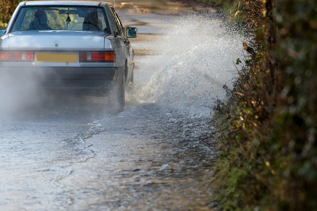 A silver car drives through a large puddle, splashing water along a narrow rural road, illustrating the impact of wet weather on vehicle surfaces.