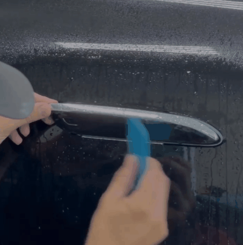 A close-up of a technician carefully applying paint protection film (PPF) to a car door handle on a black vehicle. The film is smoothed into place with a squeegee, ensuring a precise and protective fit against scratches and wear.