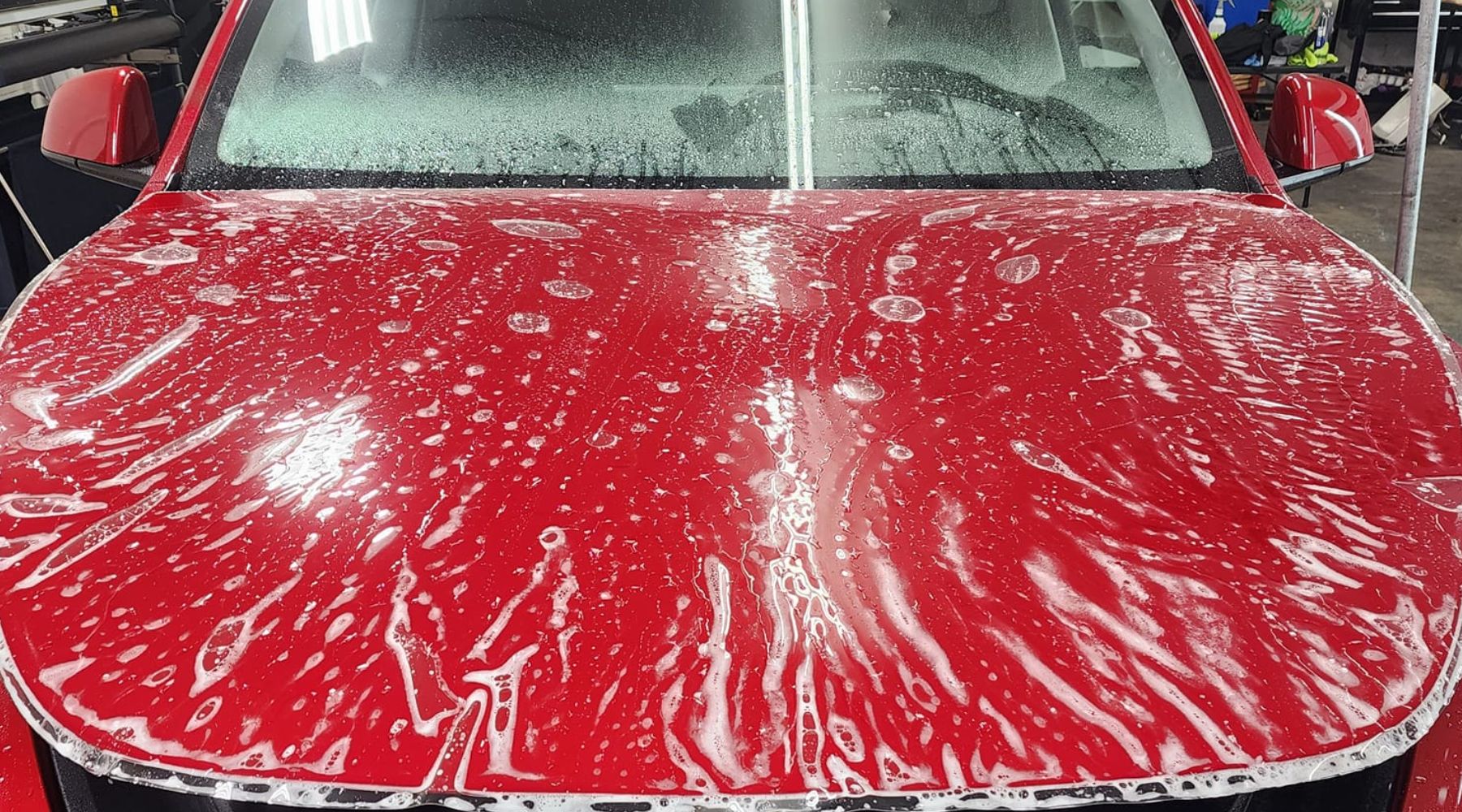 A red car's hood with paint protection film (PPF) being applied, covered in soapy water to aid in the installation process. The windshield is partially visible, and the bright indoor lig