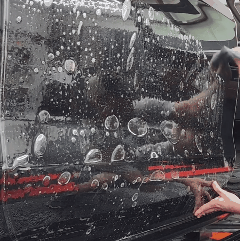 A close-up of a paint protection film (PPF) application on a black car door, with soapy water and bubbles visible as a technician smooths the film into place. The glossy surface reflects the surroundings, showcasing the film's transparency and precise installation process.