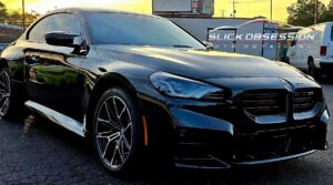 A sleek black BMW sedan with tinted windows and shiny alloy wheels, parked outside at sunset near an auto detailing shop named "Slick Obsession." The glossy finish reflects the surroundings, enhancing the vehicle's polished appearance.