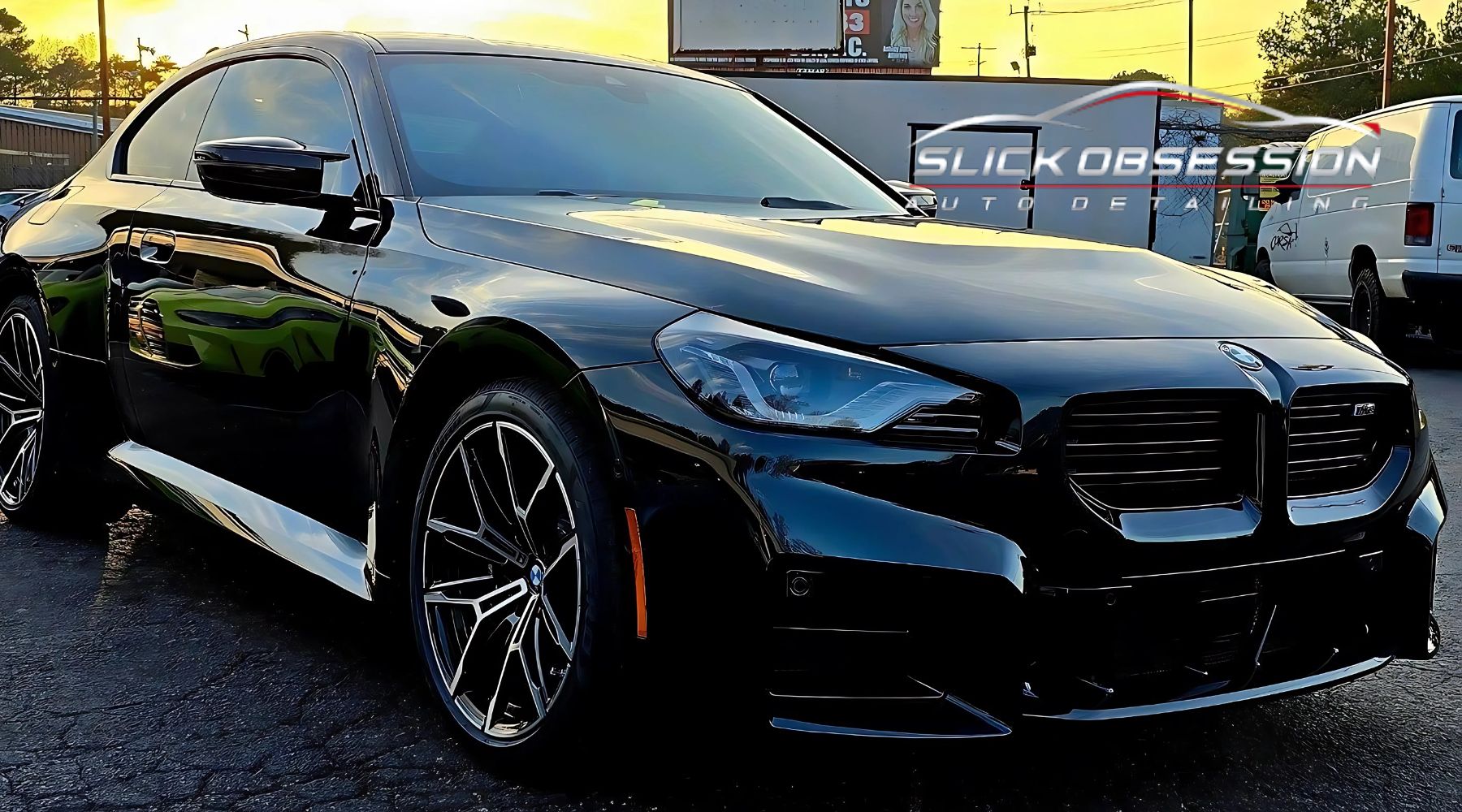 A sleek black BMW sedan with tinted windows and shiny alloy wheels, parked outside at sunset near an auto detailing shop named "Slick Obsession." The glossy finish reflects the surroundings, enhancing the vehicle's polished appearance.