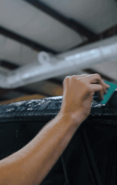 A professional installer applying paint protection film (PPF) to the roof of a black vehicle using a squeegee tool. The technician carefully smooths out the film, ensuring a bubble-free and precise application while the surface remains wet for better adhesion.