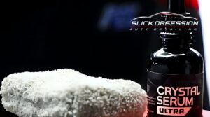 A bottle of "Crystal Serum Ultra" ceramic coating next to a soft microfiber applicator, set against a dark background. The logo for "Slick Obsession Auto Detailing" is displayed at the top, indicating a professional detailing service.
