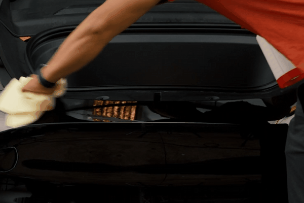 A professional detailer wiping down a glossy black car surface with a microfiber cloth, ensuring a clean and polished finish before applying paint protection film (PPF). The car's trunk area is open, and the technician's careful attention to detail highlights the precision of the PPF installation process.