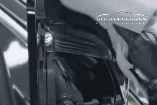A detailer applies ceramic coating to a black car’s glossy surface, using a microfiber cloth to spread the protective layer evenly. The reflection on the car’s paint becomes more pronounced as the coating is buffed in. The "Slick Obsession Auto Detailing" logo is visible in the top right corner.