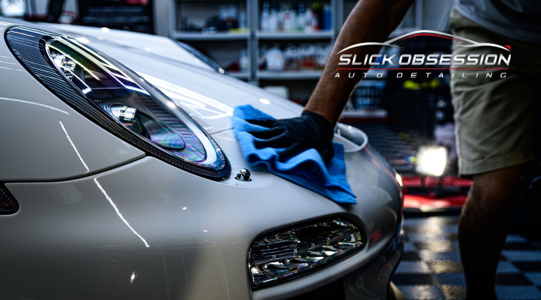 A detailer wearing black gloves uses a blue microfiber cloth to polish the glossy white hood of a sports car in a professional auto detailing shop. The reflection of lights enhances the car's pristine finish. The "Slick Obsession Auto Detailing" logo is visible in the background.
