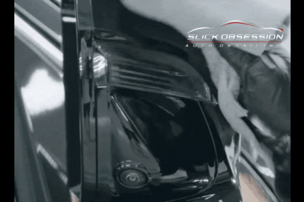A detailing technician in gloves carefully polishes a glossy black vehicle surface near the rearview camera, with the Slick Obsession Auto Detailing logo overlaid in the corner, emphasizing precision in ceramic coating removal.