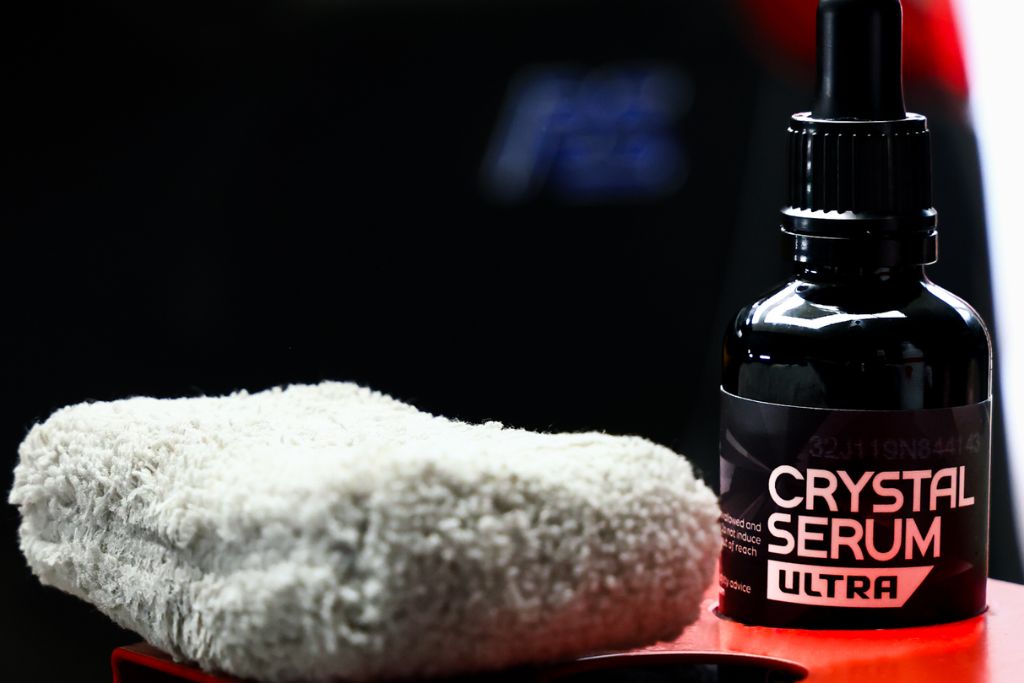Close-up of a Gtechniq Crystal Serum Ultra bottle next to a folded microfiber cloth, set against a dark backdrop with hints of a red car, highlighting tools used for professional ceramic coating application.