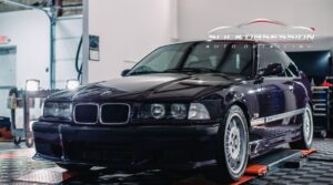 A dark BMW E36 coupe with glossy paint, tinted windows, and polished wheels inside Slick Obsession Auto Detailing shop, highlighting a professional detailing and window tint service.