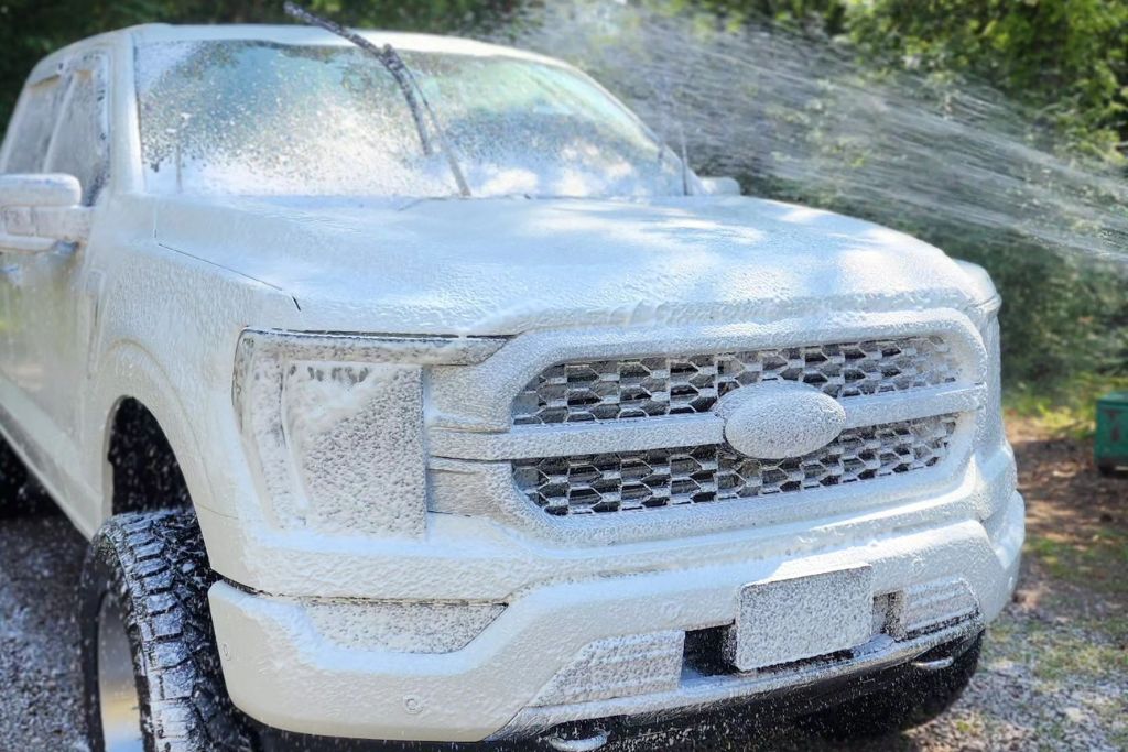 A white Ford pickup truck is fully coated in thick white foam during a wash, with water spraying over the hood and windshield. The scene emphasizes gentle cleaning practices essential for maintaining Paint Protection Film (PPF).