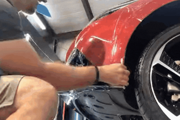 A detailing technician applies clear Paint Protection Film (PPF) to the curved front bumper of a red sports car, smoothing the transparent layer over the surface to shield it from damage. The film is visibly flexible and adheres closely to the car's contours.