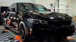 A sleek black BMW sedan with glossy paint is parked indoors at a detailing shop, reflecting lights and colors off its polished surface. The car appears freshly cleaned and coated, positioned on a checkered floor with red platforms under each wheel, highlighting a professional detailing environment.