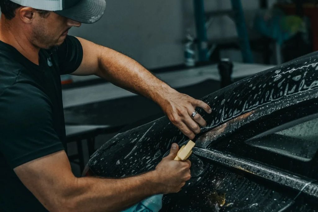 A man wearing a cap and black shirt uses a plastic scraper to remove residue from the soapy surface of a black car, focusing on the rear quarter panel near the window.