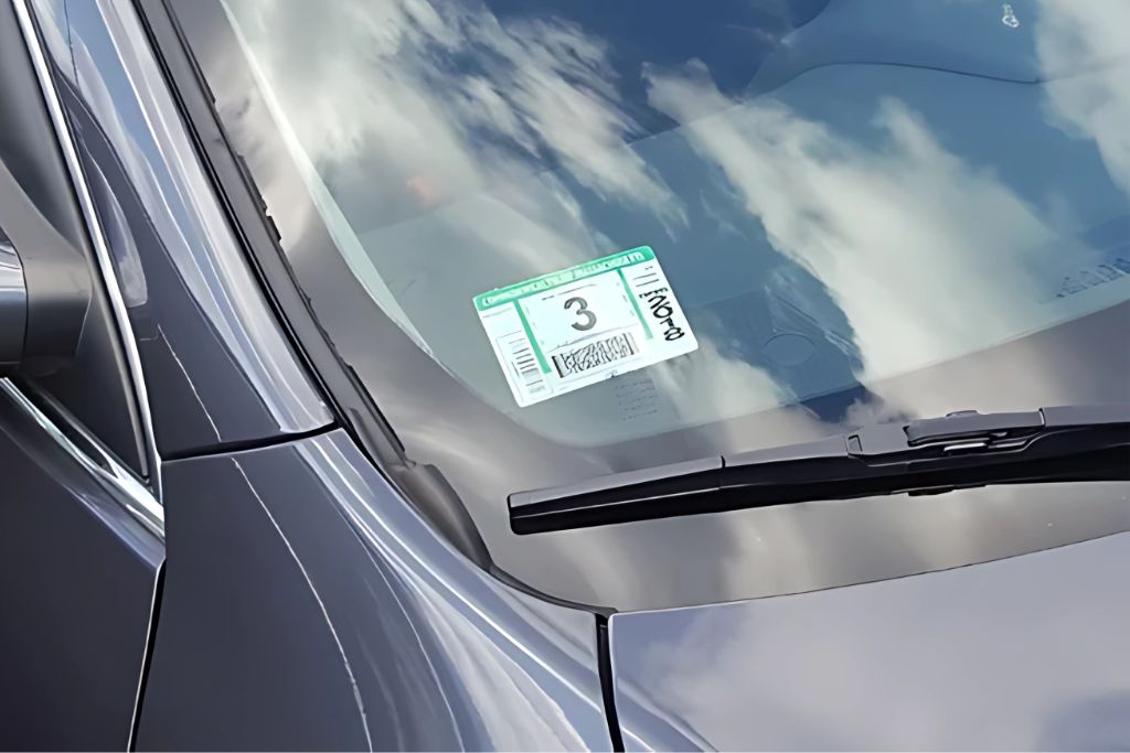 A close-up of a car windshield with a green Alabama window tint compliance sticker placed between the glass and the tint film. The surrounding reflection of clouds highlights the clarity of the tint while drawing attention to the legal documentation.