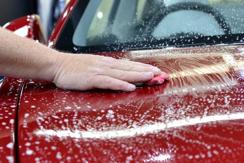Close-up of a person using a red clay bar on a wet, soapy red car hood, demonstrating a step in the DIY ceramic coating removal process.