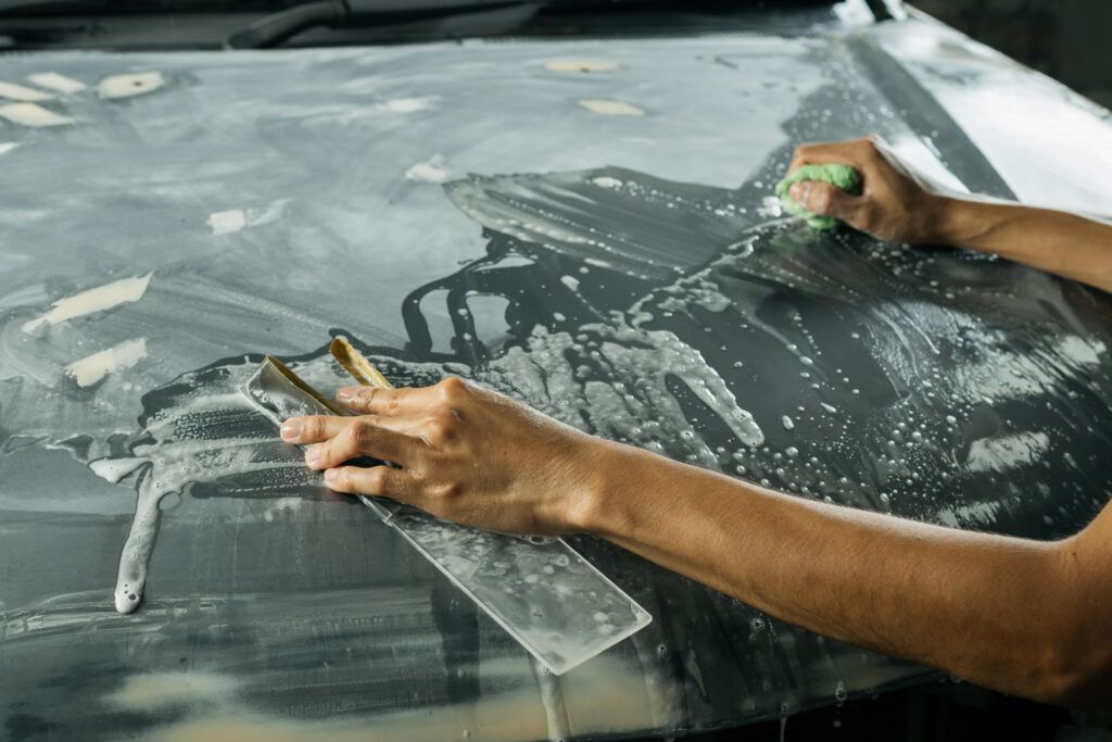 Close-up of a person using a sanding block and a green sponge to wet sand the soapy hood of a car, removing surface imperfections and high spots from a ceramic coating.