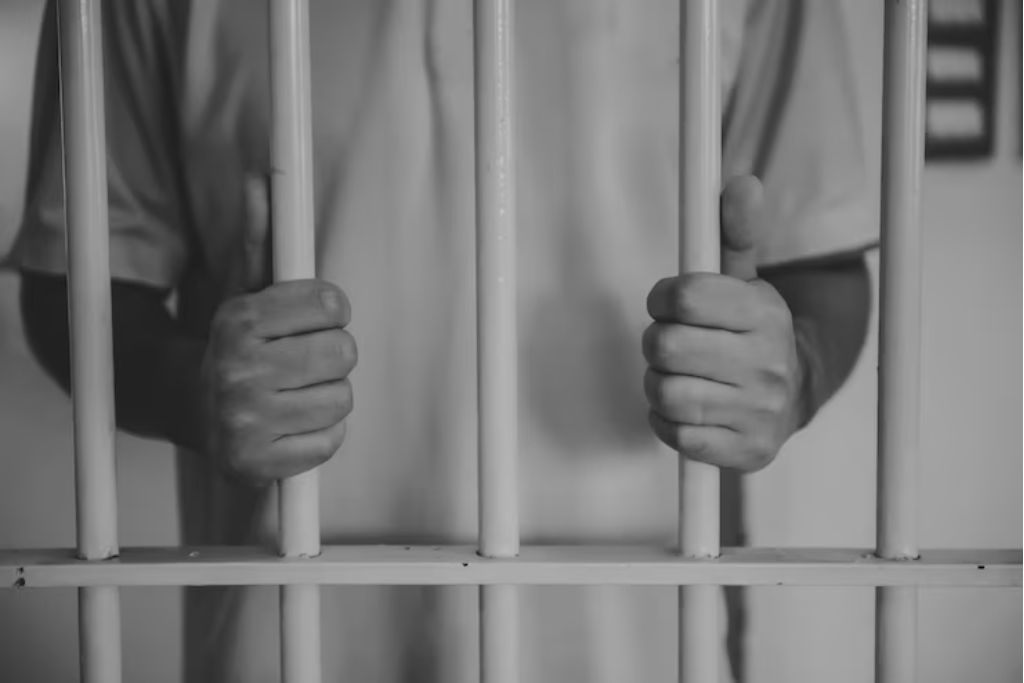 Black-and-white image showing a person gripping jail cell bars with both hands, symbolizing legal consequences. The person wears a plain shirt, and the setting conveys confinement and seriousness.