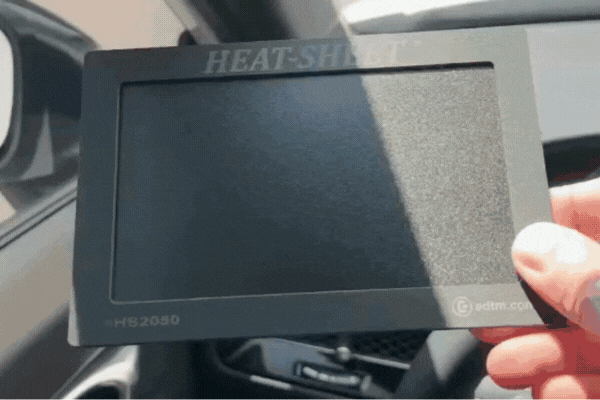A hand holds up a heat-reflecting window tint demo card labeled "HEAT-SHIELD" inside a car. As sunlight passes through the card, the tinted area visibly reduces glare and brightness, demonstrating the film’s infrared and UV-blocking capabilities.