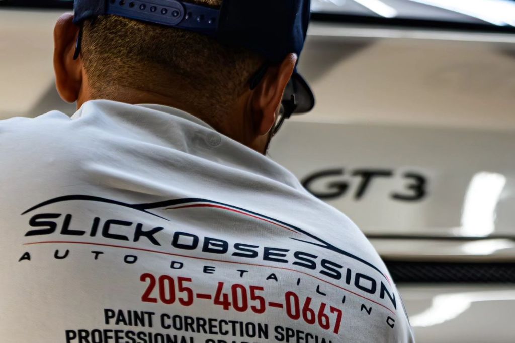 Back view of a man wearing a white “Slick Obsession Auto Detailing” shirt with a contact number and service description, standing in front of a white Porsche GT3.