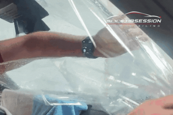 A technician carefully applies a clear window tint film to a car’s interior glass, using both hands to smooth it into place. The Slick Obsession Auto Detailing logo appears in the top corner, indicating a professional, compliant installation.