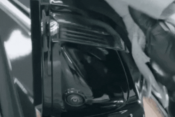 Close-up GIF of a detailer wearing black gloves using a microfiber towel to wipe down the taillight and rear panel of a black vehicle, revealing a glossy, polished surface underneath.