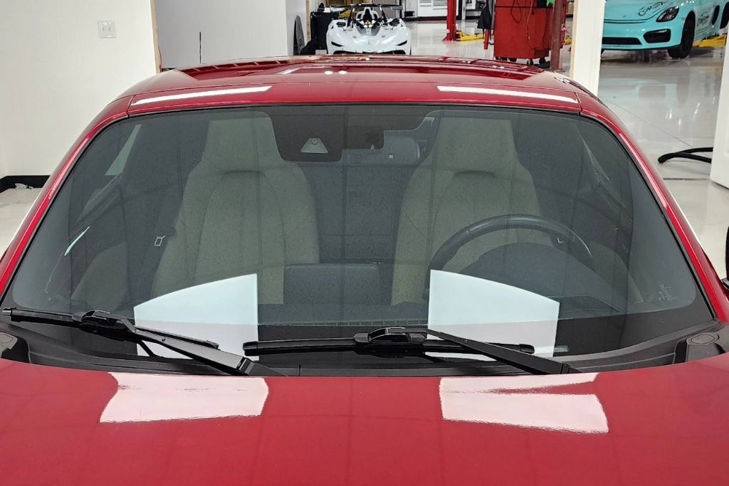 A front view of a red sports car shows a lightly tinted windshield with visible seats and dashboard inside, parked in a clean, well-lit garage. Sheets of paper placed behind the glass help demonstrate the window’s transparency, referencing legal VLT (Visible Light Transmission) standards.
