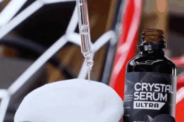 Close-up animation of a dropper applying clear ceramic coating liquid from a Crystal Serum Ultra bottle onto a soft white applicator pad, with a glossy red and black car in the blurred background.