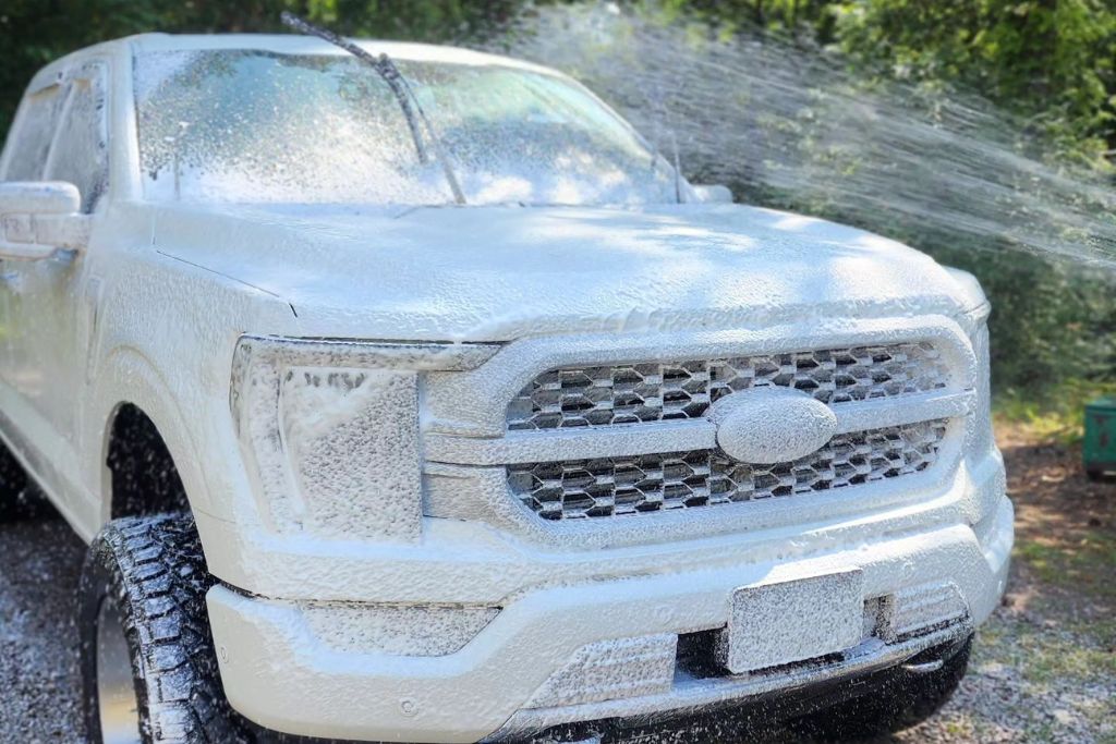 A white Ford pickup truck is being sprayed with water while covered in thick foam during a decontamination wash. The truck is parked outdoors on gravel near trees, and the foam evenly coats the entire front end, signaling a pre-coating prep process.
