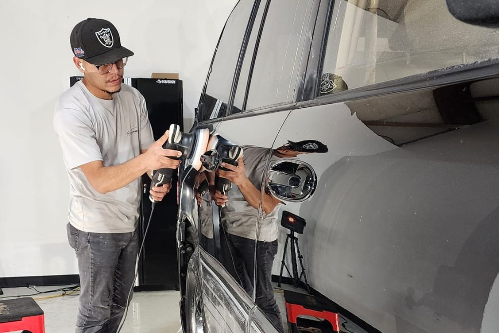 A man wearing a baseball cap and glasses uses a dual-action polisher on the side of a black SUV in a detailing shop. The glossy surface of the vehicle reflects his image clearly, highlighting the shine achieved through paint correction or ceramic coating prep.