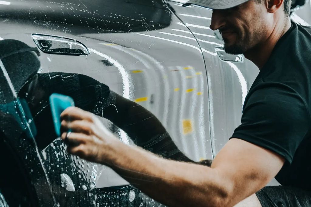 A man in a black shirt and cap applies Paint Protection Film to a glossy black car using a blue applicator, with soap and water on the surface. This process shows how PPF creates a barrier to protect the car's paint from contaminants like bird droppings, especially in sunny regions like Birmingham.