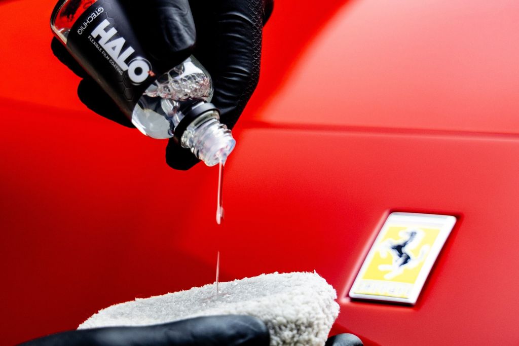 A gloved hand pours ceramic coating solution labeled “HALO” onto an applicator pad in front of a red Ferrari badge, illustrating the beginning of a professional ceramic coating process and highlighting why ceramic coating is worth it.