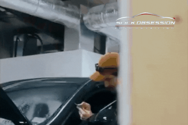 A technician in an orange cap and headlamp carefully applies Paint Protection Film to a black vehicle inside a detailing shop, with the Slick Obsession Auto Detailing logo visible. This scene reflects expert PPF installation in Birmingham, showcasing precision and care in protecting cars from bird droppings and environmental damage.