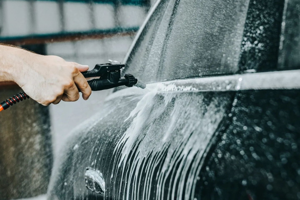 Close-up of a person using a pressure washer to clean a black vehicle covered in soap suds, focusing on maintaining a clean surface over the rear panel and window.