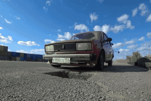 A low-angle view of an older car driving over a deep pothole on a cracked road, with the undercarriage nearly scraping the asphalt. Loose gravel and rough pavement highlight the harsh road conditions under a clear blue sky.