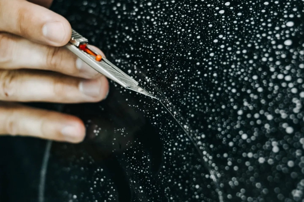 Close-up of a hand using a precision blade to trim paint protection film on a water-speckled black vehicle panel. The droplets and detail emphasize the careful installation process needed to ensure PPF performance in Birmingham’s cooler fall temperatures.
