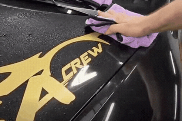 A hand wipes a glossy black hood with a purple microfiber towel, revealing a clean, hydrophobic surface with a gold “CREW” decal as water beads off. Demonstrates what is ceramic coating and how it repels water for superior paint protection.