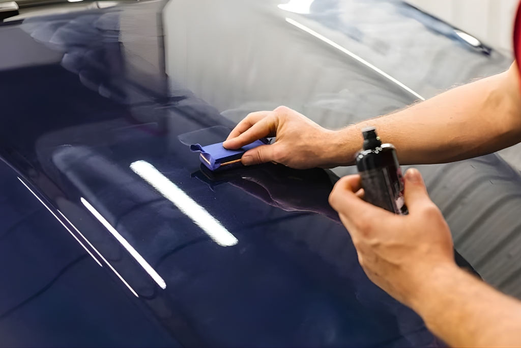 A person applies a ceramic coating with an applicator pad to a deep blue car hood inside a garage, demonstrating a DIY kit process. This image supports the comparison between professional and at-home applications in the context of DIY Kits for ceramic coating in Birmingham, AL.