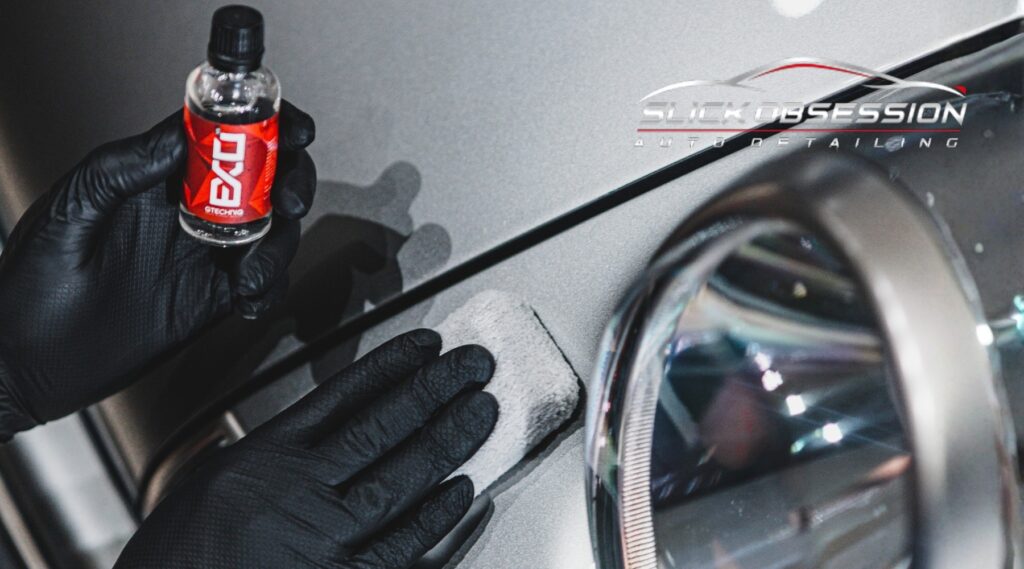 A close-up of a hand wearing a black glove holding a Gtechniq EXO ceramic coating bottle, applying the product with a microfiber pad to a metallic silver vehicle’s body near the headlight, with the Slick Obsession Auto Detailing logo. This image visually supports the topic "What Is Ceramic Coating" as demonstrated by Birmingham's Slick Obsession Auto Detailing.