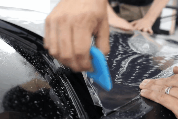 Close-up of a technician applying paint protection film to the hood of a black vehicle using a blue squeegee, with water and film visible during the smoothing process. This shows the precision involved in partial PPF coverage, commonly chosen for budget-conscious drivers in Birmingham, AL.