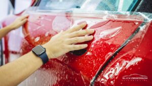 A person smooths out Paint Protection Film on a red car door using their hands, with water droplets still present on the glossy surface.