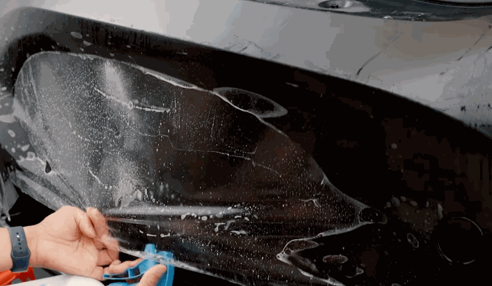 Close-up of a person applying Paint Protection Film to a wet black car surface using a blue squeegee, with water droplets and air bubbles visible under the film.