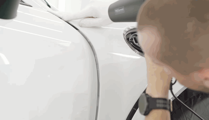 Detailer using a heat gun to set the paint protection film along the curved edge of a white car’s fender.