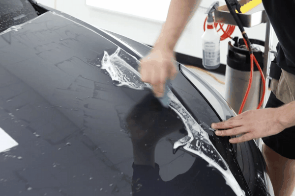 Technician using a squeegee to apply paint protection film on a car hood covered in slip solution, smoothing out bubbles during installation. This close up shows the hands on process that affects how long PPF installation takes depending on coverage and curing time.