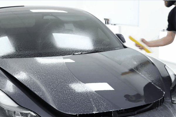 Technician applying paint protection film to the full front of a black vehicle using a yellow squeegee while the hood and windshield are covered in installation solution. The image represents Full Front and Track Pack PPF coverage in Birmingham AL, highlighting protection for the hood, bumper, fenders, headlights, and high impact driving areas.