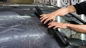 Alt text. Technician applying Ceramic Window Tint film to a car windshield using a squeegee to smooth out water and air bubbles during installation inside an auto detailing shop.