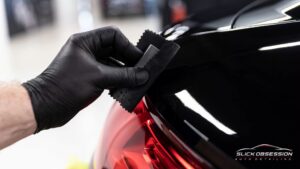 Close up of a gloved hand applying ceramic coating near a red tail light on a black vehicle to protect the paint and deepen gloss.