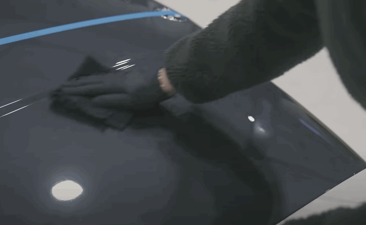 Gloved hand leveling ceramic coating on a dark car hood with a microfiber cloth to create a smooth high gloss finish.