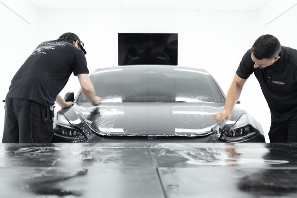 Two professional installers applying paint protection film across the full front of a car inside a clean indoor detailing bay, smoothing the film over the hood and headlights. The image supports why choosing professional PPF installers ensures proper application, controlled conditions, and long lasting results compared to DIY installation.