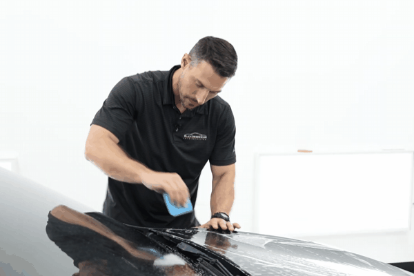 Professional technician using a blue squeegee to install paint protection film on a glossy black vehicle hood inside a clean controlled detailing facility. The image highlights the precision required during PPF installation to prevent trapping scratches or defects under the film.